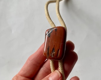 May include: A bolo tie featuring a rectangular, polished, reddish-brown stone with black veining. The stone is suspended from a beige cord. The cord is held in a hand.
