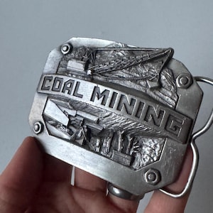 Könnte beinhalten: Silberfarbene Gürtelschnalle mit dem geprägten Schriftzug "COAL MINING" in der Mitte. Die Schnalle zeigt ein detailliertes Relief einer Kohlebergbauszene, einschließlich Maschinen und Strukturen. Die Schnalle ist rechteckig mit abgerundeten Ecken.