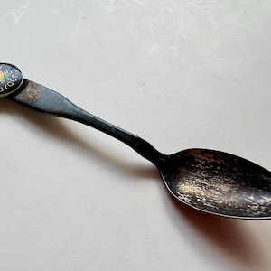 May include: A vintage silver spoon with a black handle and bowl. The spoon has a small, round emblem attached to the handle, featuring a yellow rose and the words "Minter Gardens" in white text on a black background.