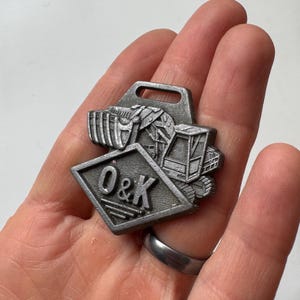 May include: A silver-colored metal pin featuring a construction vehicle design. The pin includes the letters "O&K" within a diamond shape and a rectangular top with a hole.