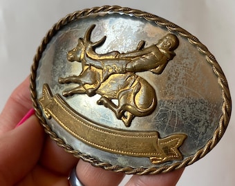May include: Oval-shaped silver and gold-tone belt buckle with a cowboy riding a bucking bronco. The buckle has a twisted rope border and a banner at the bottom. A classic Western-themed accessory.