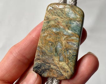 May include: A rectangular bolo tie pendant with a polished, multicolored stone. The stone features shades of green, brown, and blue with a marbled pattern. The pendant is attached to a light gray braided cord.