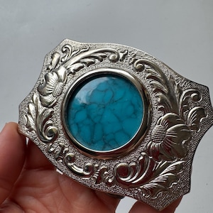 May include: Silver-toned belt buckle with a turquoise-colored stone center. The buckle has an ornate floral and scroll design around the stone. The buckle is rectangular with rounded corners.