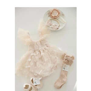 May include: A beige baby outfit with a tulle skirt and butterfly accents. The outfit includes a headband with a flower, a pair of beige socks with a bow, and a pair of beige baby shoes with butterfly accents.