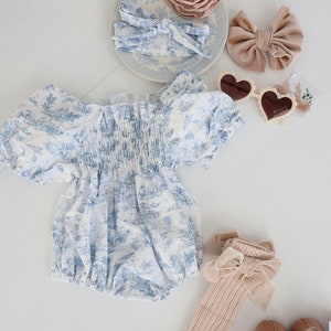 May include: A baby outfit set with a white and blue floral print romper, a pair of beige socks with a brown velvet bow, a pair of brown mary jane shoes, a brown velvet bow headband, a white headband with a blue floral print, a beige flower hair clip, and a pair of heart-shaped sunglasses.