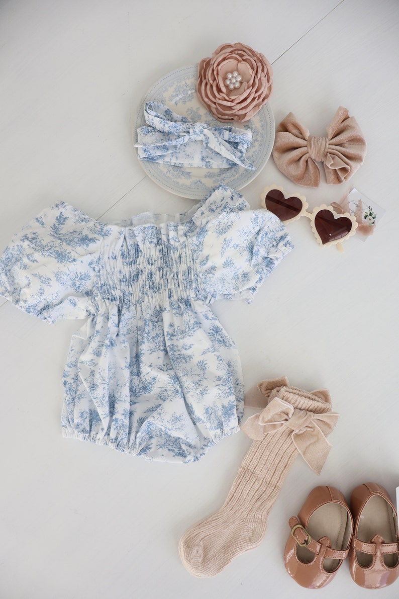 May include: A baby outfit set with a white and blue floral print romper, a pair of beige socks with a brown velvet bow, a pair of brown mary jane shoes, a brown velvet bow headband, a white headband with a blue floral print, a beige flower hair clip, and a pair of heart-shaped sunglasses.