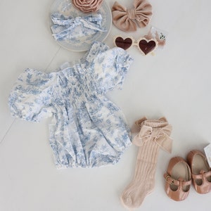 May include: A baby outfit set with a blue and white floral print romper, a pair of beige socks with a brown bow, a pair of brown shoes, a brown velvet bow, a white headband with a blue and white floral print, and a brown flower hair clip.