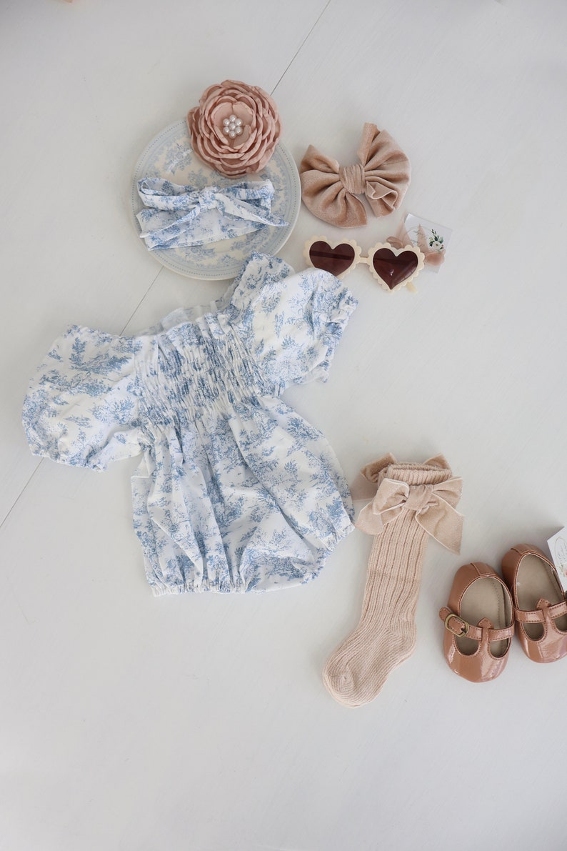 May include: A baby outfit set with a blue and white floral print romper, a pair of beige socks with a brown bow, a pair of brown shoes, a brown velvet bow, a white headband with a blue and white floral print, and a brown flower hair clip.