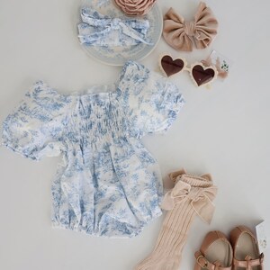 May include: A baby outfit set with a blue and white floral print romper, a pair of beige socks with a brown bow, a pair of brown shoes with a buckle, a headband with a brown bow, a headband with a pink flower, and a pair of heart-shaped sunglasses.