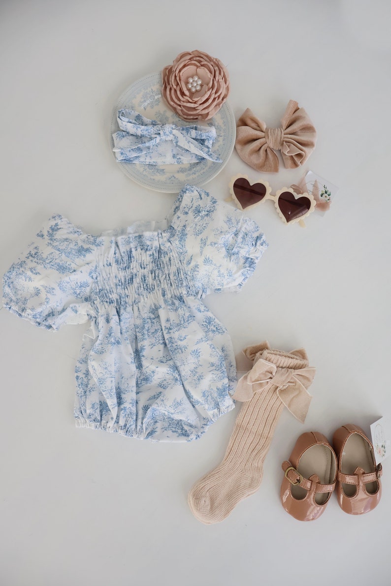 May include: A baby outfit set with a blue and white floral print romper, a pair of beige socks with a brown bow, a pair of brown shoes with a buckle, a headband with a brown bow, a headband with a pink flower, and a pair of heart-shaped sunglasses.