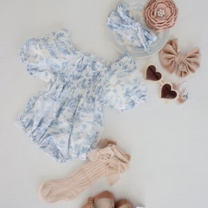 May include: A baby outfit set with a blue and white floral print dress, a pair of beige socks with a bow, a pair of brown shoes, a beige headband with a bow, a beige flower hair clip, and a pair of heart-shaped sunglasses.