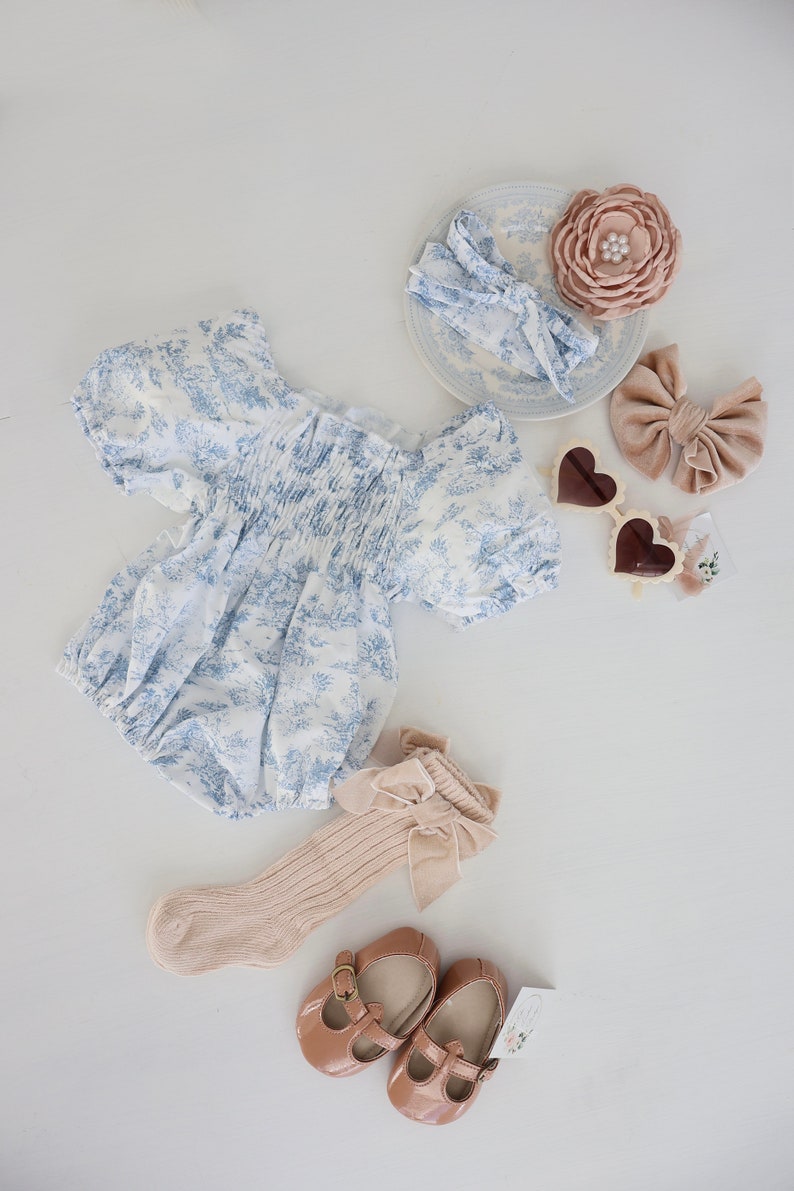May include: A baby outfit set with a blue and white floral print dress, a pair of beige socks with a bow, a pair of brown shoes, a beige headband with a bow, a beige flower hair clip, and a pair of heart-shaped sunglasses.