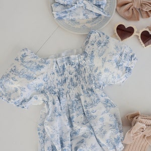 May include: A white and blue floral print baby onesie with puffed sleeves and a gathered elastic waistband. The onesie is laid out on a white surface with a brown velvet bow, a white headband with a blue floral print, a pink flower, and a pair of heart-shaped sunglasses.