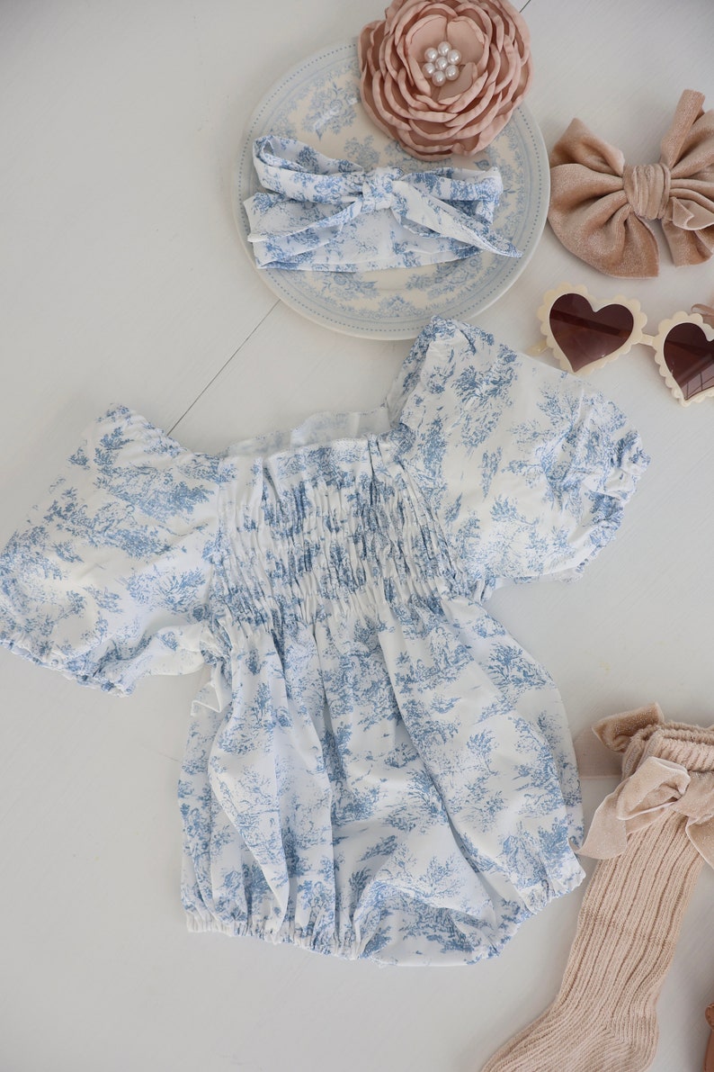 May include: A white and blue floral print baby onesie with puffed sleeves and a gathered elastic waistband. The onesie is laid out on a white surface with a brown velvet bow, a white headband with a blue floral print, a pink flower, and a pair of heart-shaped sunglasses.