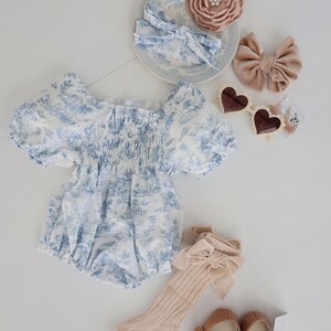 May include: A baby outfit set with a blue and white floral print romper, a pair of beige socks with a brown bow, a pair of brown mary jane shoes, and two hair bows. One hair bow is brown velvet and the other is a floral print headband.