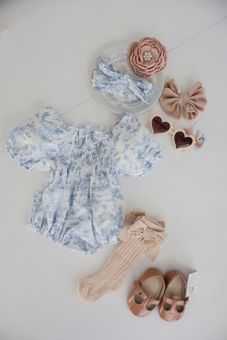 May include: A baby outfit set with a blue and white floral print romper, a pair of beige socks with a brown bow, a pair of brown mary jane shoes, and two hair bows. One hair bow is brown velvet and the other is a floral print headband.