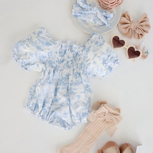 May include: A blue and white floral print baby onesie with puffed sleeves, a pair of beige socks with a brown velvet bow, a pair of brown leather mary jane shoes, a pair of white heart-shaped sunglasses, a blue and white headband, and a pink flower headband.