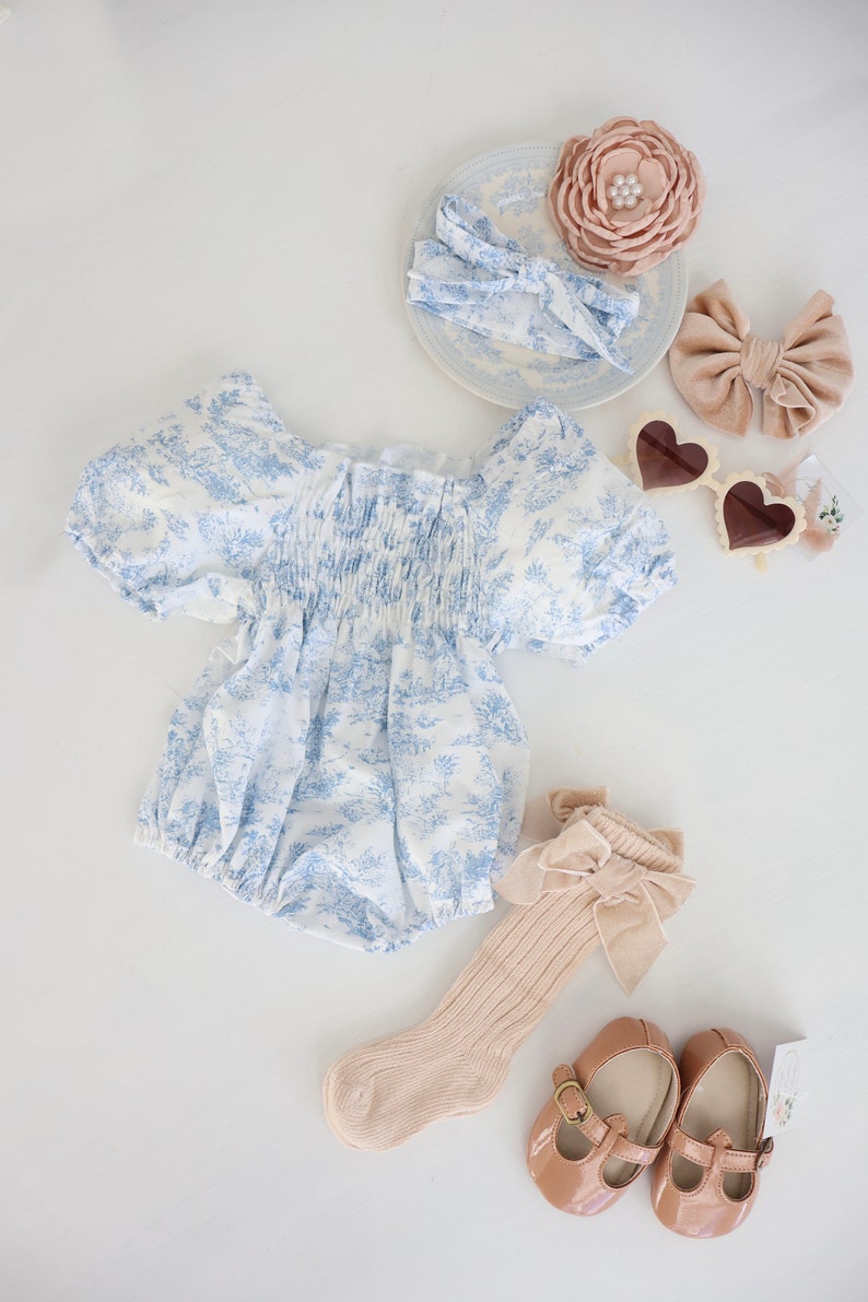 May include: A blue and white floral print baby onesie with puffed sleeves, a pair of beige socks with a brown velvet bow, a pair of brown leather mary jane shoes, a pair of white heart-shaped sunglasses, a blue and white headband, and a pink flower headband.