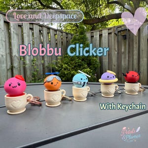 May include: A collection of five colorful "Blobbu Clicker" keychains, each featuring a unique character atop a small teacup. The keychains are in various colors, including pink, orange, blue, purple, and red, with attached key rings. The text "Love and Deepspace" and "Petals & Plumes" are also visible.