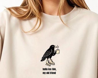 May include: Sand-colored sweatshirt with a graphic of a black crow on ice skates holding a mug. The text "hello ice rink, my old friend" is below the graphic. The sweatshirt is a neutral color.