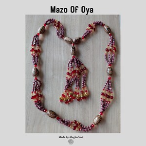 Può includere: Una collana Mazo of Oya con un motivo ripetitivo di perline color marrone scuro e giallo, intervallate da perline più grandi di forma ovale con macchie marroni e bianche. La collana include una nappa centrale fatta di fili di perline.