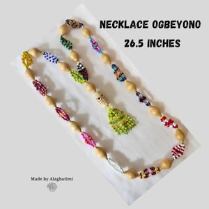 Könnte beinhalten: Eine Perlenkette mit der Aufschrift "NECKLACE OGBEYONO" ist 67,3 cm lang. Die Kette besteht aus Holzperlen, die mit farbigen Perlenabschnitten in verschiedenen Mustern durchsetzt sind. Ein Totenkopf-Anhänger mit einer gelb-grünen Quaste hängt in der Mitte.