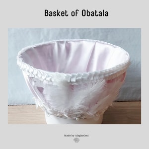 May include: A white basket featuring a satin interior lining, a pleated white ribbon trim along the top edge, and white feathers accented with small white beads. The text "Basket of Obatala" and "Made by AlagbaOmi" appears on the image.