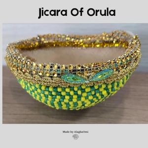 May include: A Jicara Of Orula bowl featuring a yellow and green beaded exterior. The rim is decorated with gold-toned metallic trim, small circular rhinestones, and two prominent teal-coloured marquise-shaped crystals. Text reads Jicara Of Orula.
