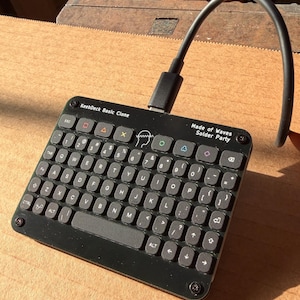 Puede incluir: Un teclado KeebDeck Basic Clone negro con un cable USB. El teclado tiene un diseño compacto con letras y símbolos en las teclas. El texto "Made of Waves Solder Party" está impreso en la parte superior derecha.