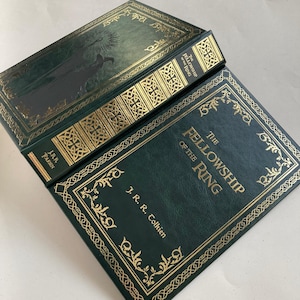 May include: A green hardcover book titled "The Fellowship of the Ring" by J.R.R. Tolkien. The book has gold lettering and decorative borders. The spine also features gold lettering. The cover has a silhouette of a figure.