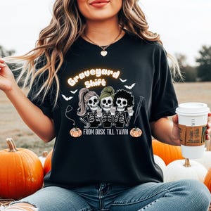 Puede incluir: Camiseta negra con un diseño temático de Halloween. El diseño presenta tres figuras de esqueleto sosteniendo tazas de café, con las palabras "Graveyard Shift" y "From Dusk Till Yawn". Incluye murciélagos y calabazas.
