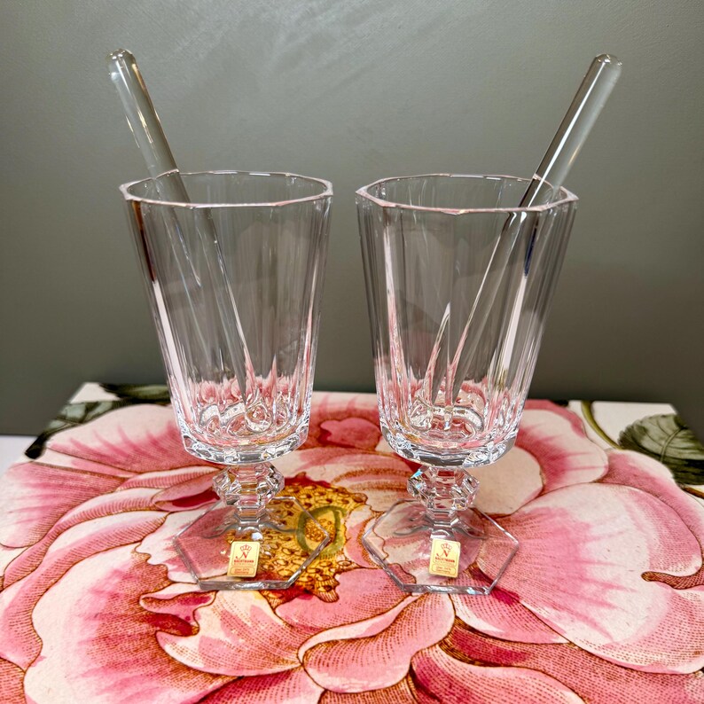 K&ouml;nnte beinhalten: Zwei klare Cocktailgl&auml;ser aus Kristall mit facettiertem Design, jeweils mit einem R&uuml;hrstab aus klarem Glas. Die Gl&auml;ser haben einen sechseckigen Fu&szlig; und einen dekorativen Stiel. Die Gl&auml;ser stehen auf einer Oberfl&auml;che mit floralem Muster.