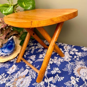 May include: A small, wooden folding stool with a light brown finish. The stool has a rounded top and four angled legs that cross in an X-shape. The stool is set on a blue floral patterned surface.