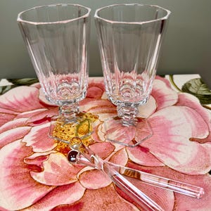 K&ouml;nnte beinhalten: Zwei klare, facettierte Kristall-Champagnerfl&ouml;ten mit sechseckigen B&ouml;den und Stielen. Zwei klare, lange, d&uuml;nne R&uuml;hrst&auml;bchen mit einer kleinen roten Kugel am Ende. Auf einer Oberfl&auml;che mit Blumenmuster.