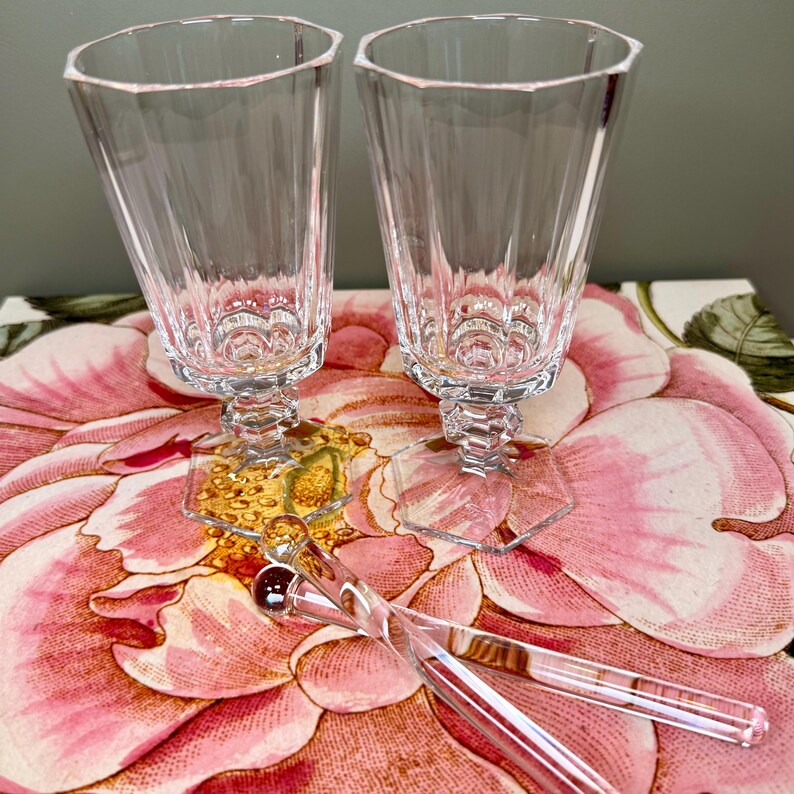 K&ouml;nnte beinhalten: Zwei klare, facettierte Kristall-Champagnerfl&ouml;ten mit sechseckigen B&ouml;den und Stielen. Zwei klare, lange, d&uuml;nne R&uuml;hrst&auml;bchen mit einer kleinen roten Kugel am Ende. Auf einer Oberfl&auml;che mit Blumenmuster.