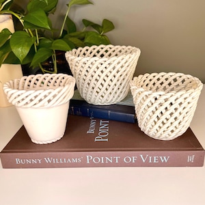 May include: Three off-white ceramic woven baskets of varying sizes. The baskets are arranged on a brown book with the title "Bunny Williams' Point of View". A green plant is visible in the background.