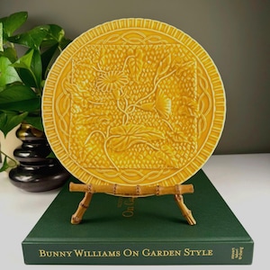 May include: A yellow ceramic plate with a floral design, displayed on a bamboo stand. The plate features a raised pattern of flowers and leaves within a square and circular border. The plate is resting on a green book titled "Bunny Williams On Garden Style."
