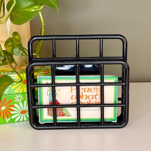 May include: Black metal recipe card holder with a grid design, holding vintage recipe cards. The cards have a green and white border with the text "Here's what's cookin". A decorative floral box is visible in the background.