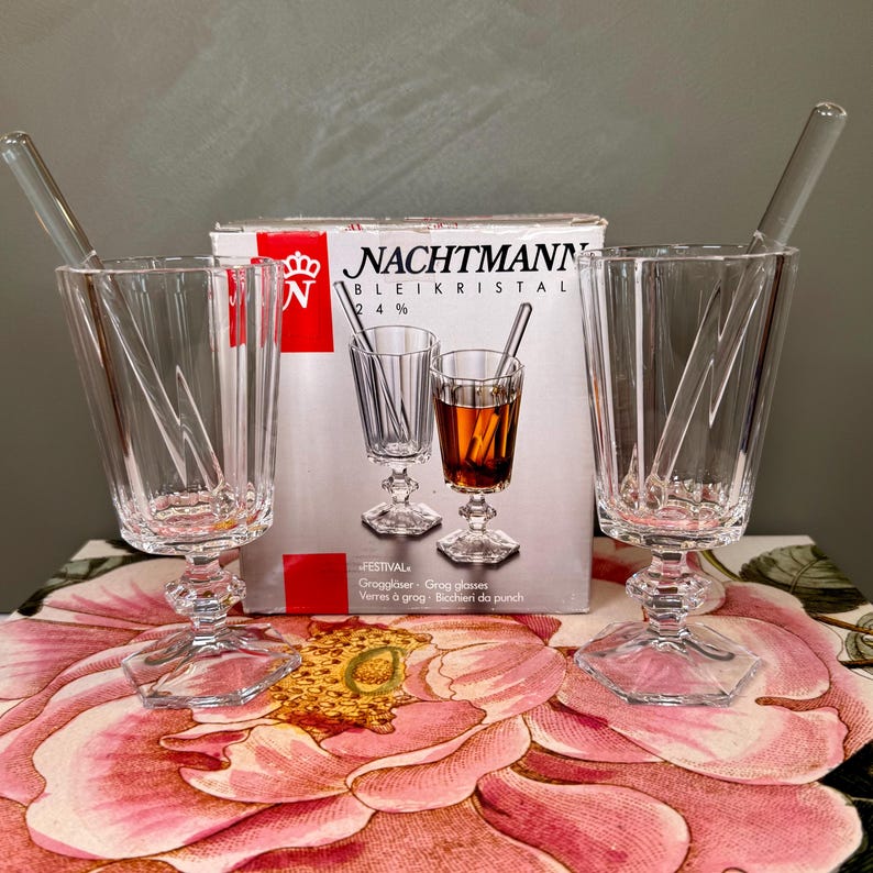 K&ouml;nnte beinhalten: Klare Kristall-Grog-Gl&auml;ser mit R&uuml;hrst&auml;bchen. Die Gl&auml;ser haben ein facettiertes Design und stehen auf einem dekorativen Sockel. Eine Schachtel mit Nachtmann Kristall-Grog-Gl&auml;sern befindet sich hinter den Gl&auml;sern.