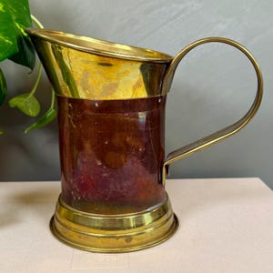 May include: Vintage copper and brass pitcher with a curved handle. The pitcher has a cylindrical body with a brass rim and base. The handle is also brass. The copper body has a reddish-brown patina.