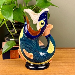May include: A ceramic fish-shaped pitcher with a wide open mouth. The pitcher is primarily blue with yellow, orange, and white accents. The fish has a detailed scale pattern and a black base. The pitcher is a decorative item.
