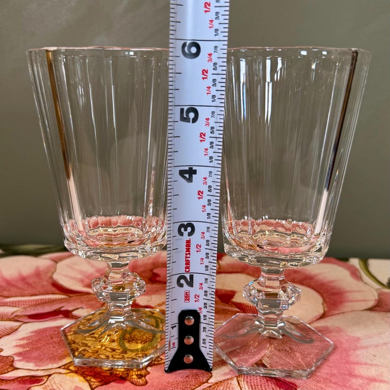 K&ouml;nnte beinhalten: Zwei klare Kristall-Weingl&auml;ser mit vertikal gerippten Seiten und sechseckigen B&ouml;den. Die Gl&auml;ser sind etwa 12.7 cm hoch und haben einen dekorativen Stiel. Ein Ma&szlig;band befindet sich hinter den Gl&auml;sern.