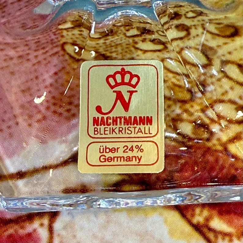 K&ouml;nnte beinhalten: Nahaufnahme eines transparenten Kristallglasobjekts mit einem gold-roten Aufkleber. Der Aufkleber lautet "NACHTMANN BLEIKRISTALL" und "&uuml;ber 24% Germany". Das Glas hat eine strukturierte Oberfl&auml;che.