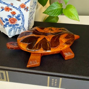 May include: A handcrafted wooden turtle figurine with a detailed leaf and floral design on its shell. The turtle is made of light and dark brown wood and has four legs and a small head. The figurine is approximately 6 inches long.