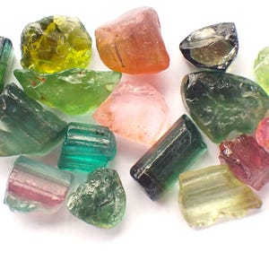 May include: A selection of raw gemstones in shades of green, pink, orange, and black. The gemstones are displayed on a white background, revealing their natural textures and forms. The image emphasises the variety of the stones.
