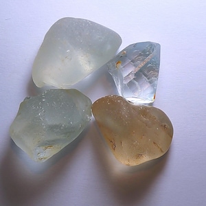 May include: Four pieces of sea glass with smooth, rounded edges. Two pieces are pale, translucent green, one is a clear, angular shard with a slight blue tint, and one is a rounded, opaque amber-coloured piece with a textured, weathered surface.