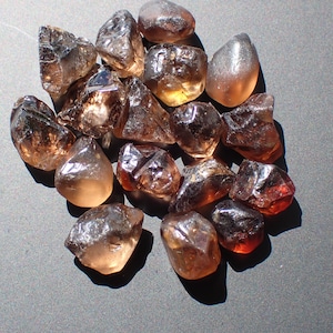 May include: A cluster of raw smoky quartz crystals. The crystals, in various sizes and shapes, exhibit amber and brown tones. The gemstones are arranged on a grey surface, with light reflecting off their facets. Measurements are in cm.