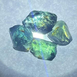 May include: A cluster of four raw, translucent gemstones with a vibrant teal and green colour palette. The stones feature irregular, angular shapes with faceted surfaces that catch the light, displaying internal golden inclusions and bright highlights.