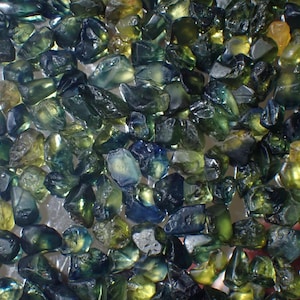 Puede incluir: Primer plano de una colección de piedras preciosas en bruto. Las piedras son una mezcla de tonos verdes, azules y amarillos, con una calidad translúcida. Las formas y tamaños irregulares de las piedras sugieren que están en su estado natural, sin pulir.