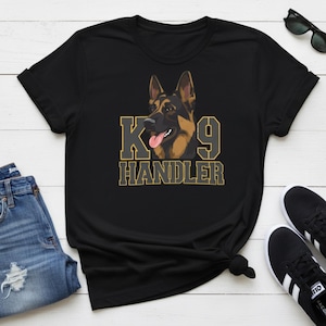 May include: Black t-shirt with a German Shepherd dog graphic and the text "K9 Handler" in gold. The dog has its tongue out. The shirt is styled with blue jeans and black trainers.